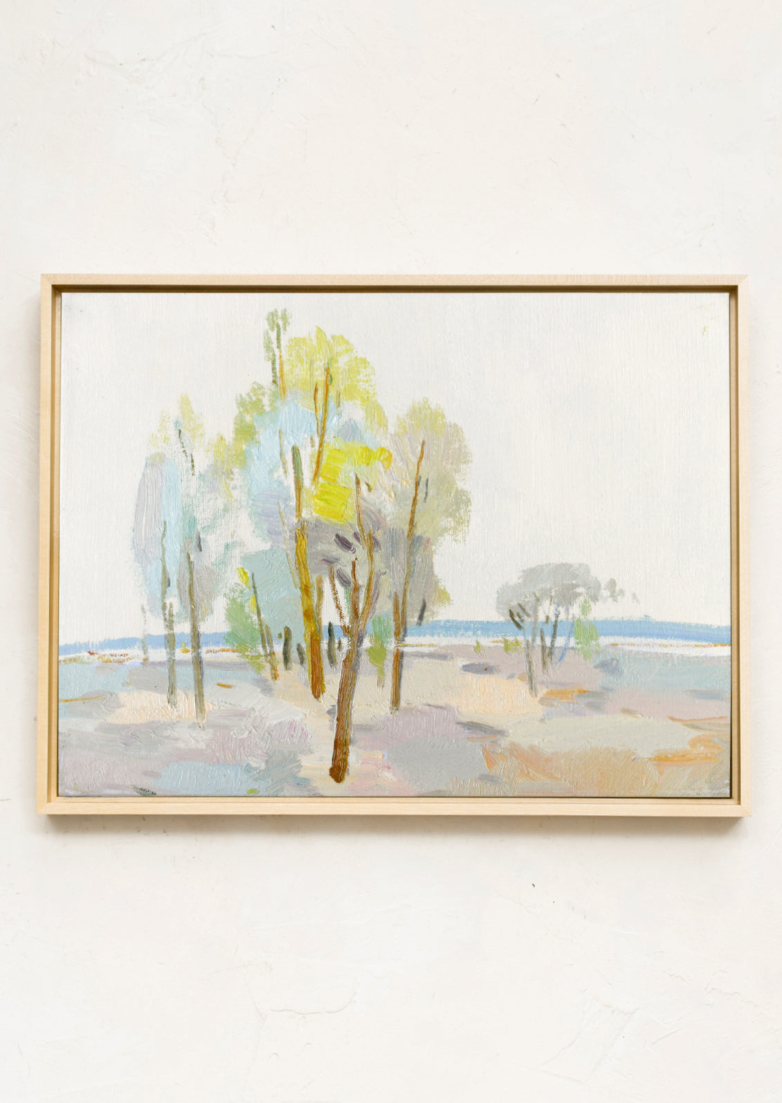 A framed landscape painting featuring stylized trees in soft pastel shades of green, blue, beige, and gray, housed in a light natural wood frame and mounted on a textured off-white wall.