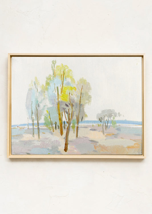 A framed landscape painting featuring stylized trees in soft pastel shades of green, blue, beige, and gray, housed in a light natural wood frame and mounted on a textured off-white wall.