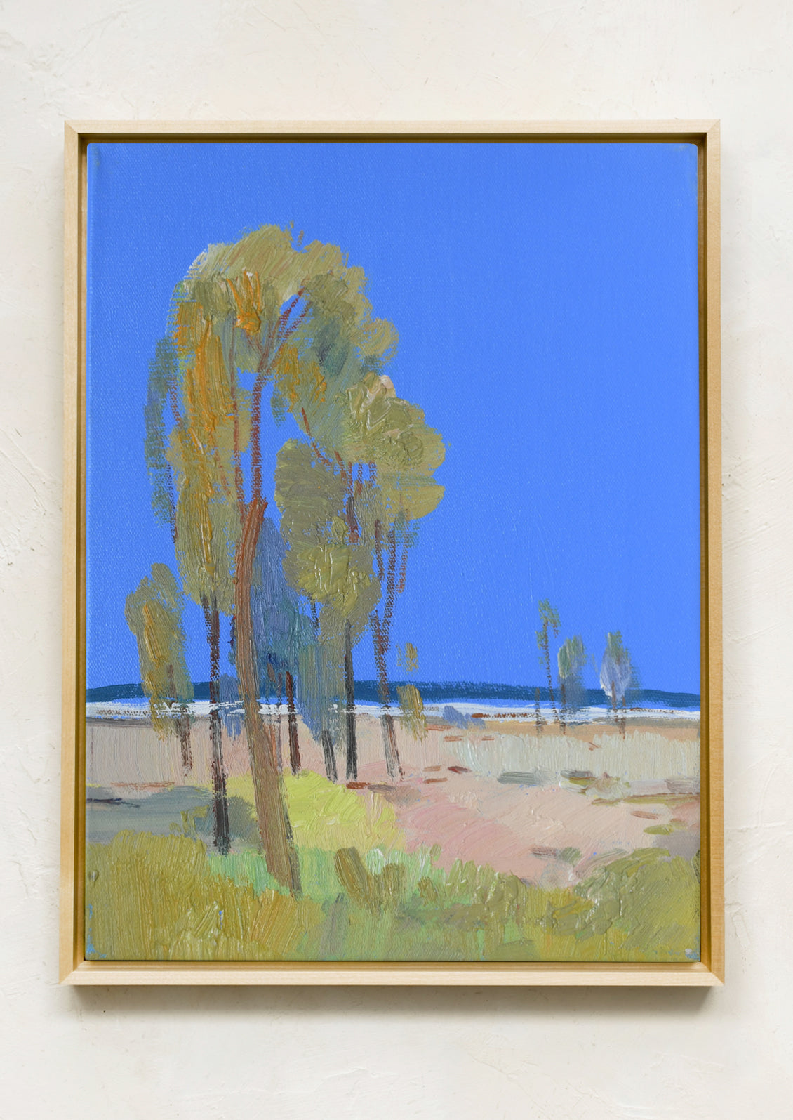 A framed landscape painting featuring tall green trees under a bright blue sky, hung on a textured off-white wall.