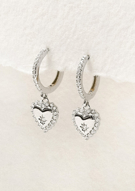 A pair of sterling silver huggie hoop earrings with dangling heart charms adorned with cubic zirconia gems.