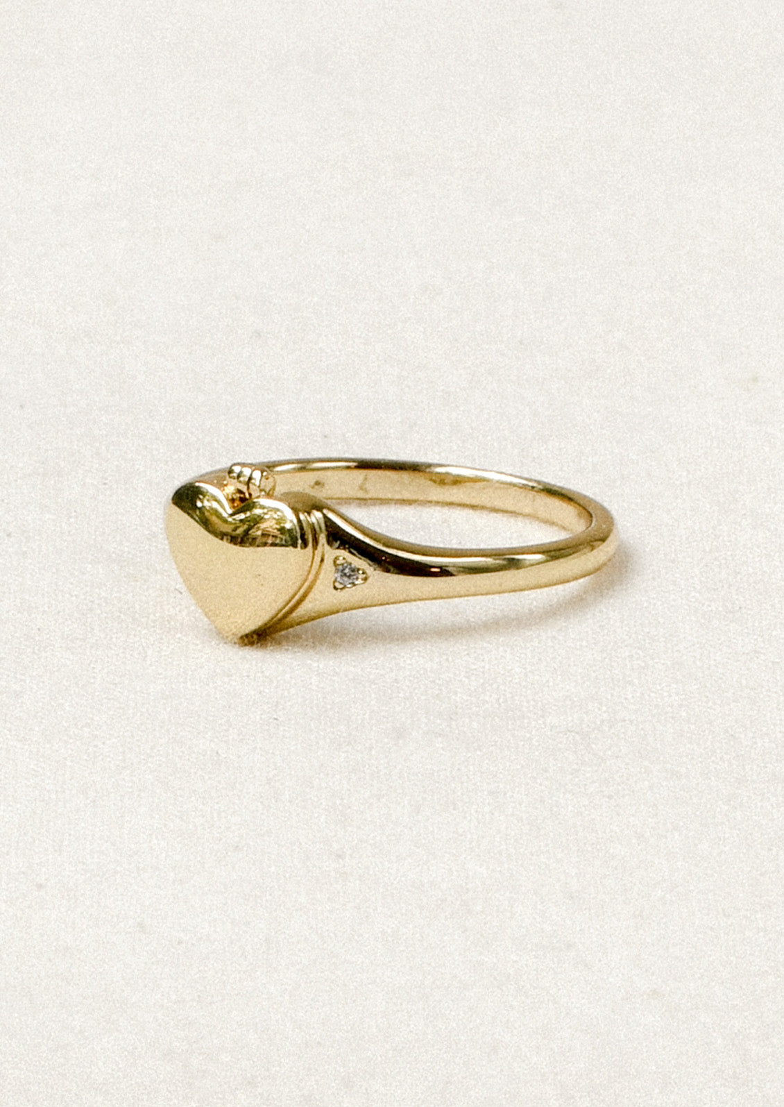 A gold ring with a tiny locket in the shape of a heart in the center and crystal detailing on either side.