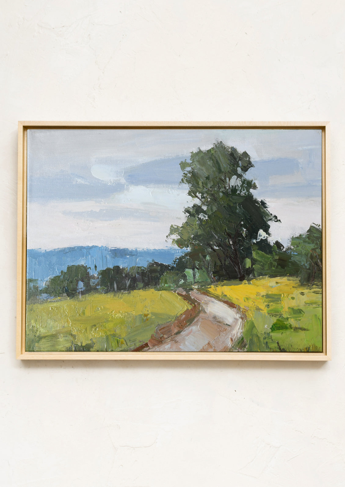 A framed landscape painting on a textured off-white wall, depicting a winding dirt path through a green field under a cloudy blue sky.