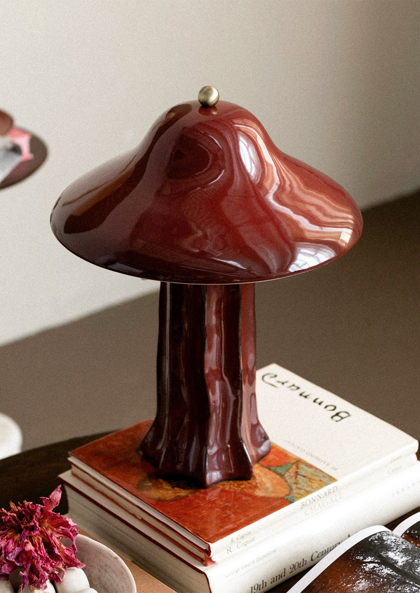 A glossy mushroom-shaped table lamp in a deep reddish-brown finish, displayed on top of stacked books in an indoor setting.