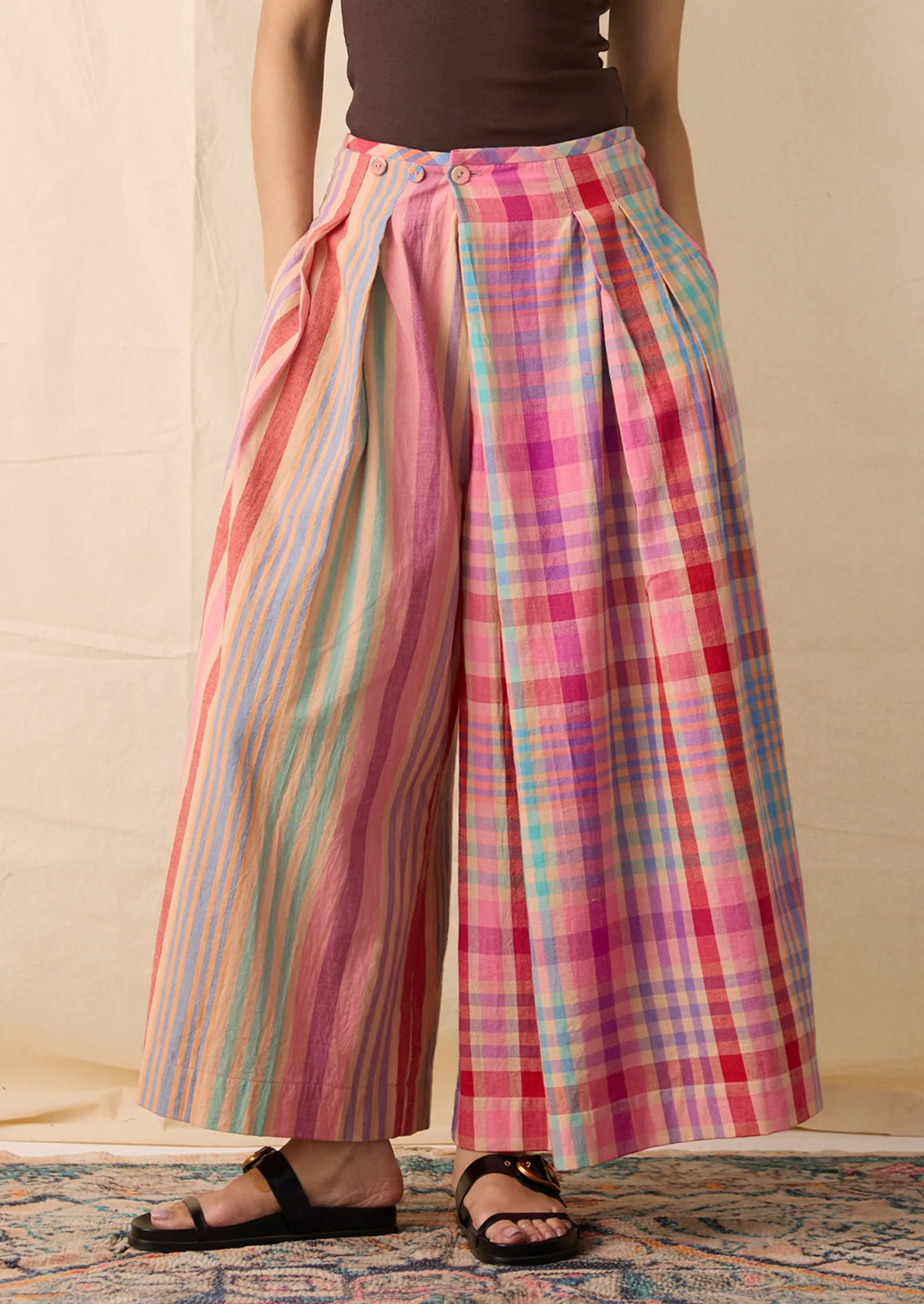 A person wearing wide-leg cropped pants with a split design: one leg has vertical pastel stripes in pink, blue, yellow, and green, and the other leg has a pink, red, and blue plaid pattern.