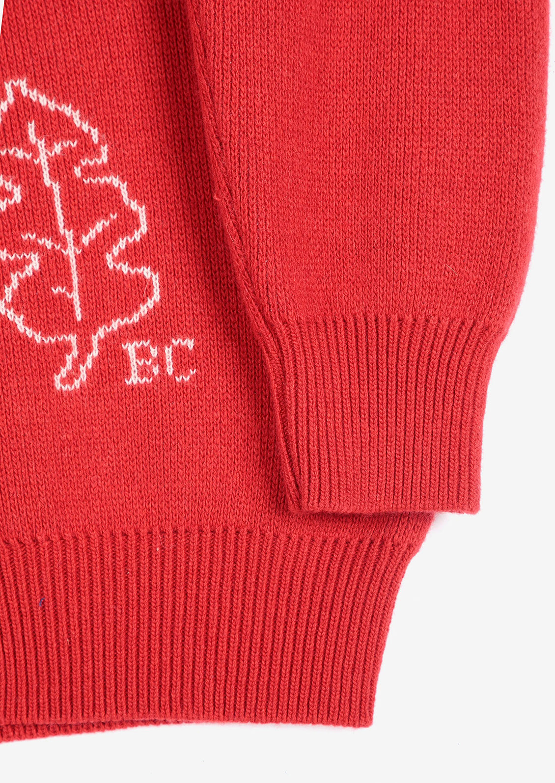 Close-up of a red knit garment showing a white embroidered leaf motif and the letters 'BC' on the chest, with ribbed cuffs and hem.