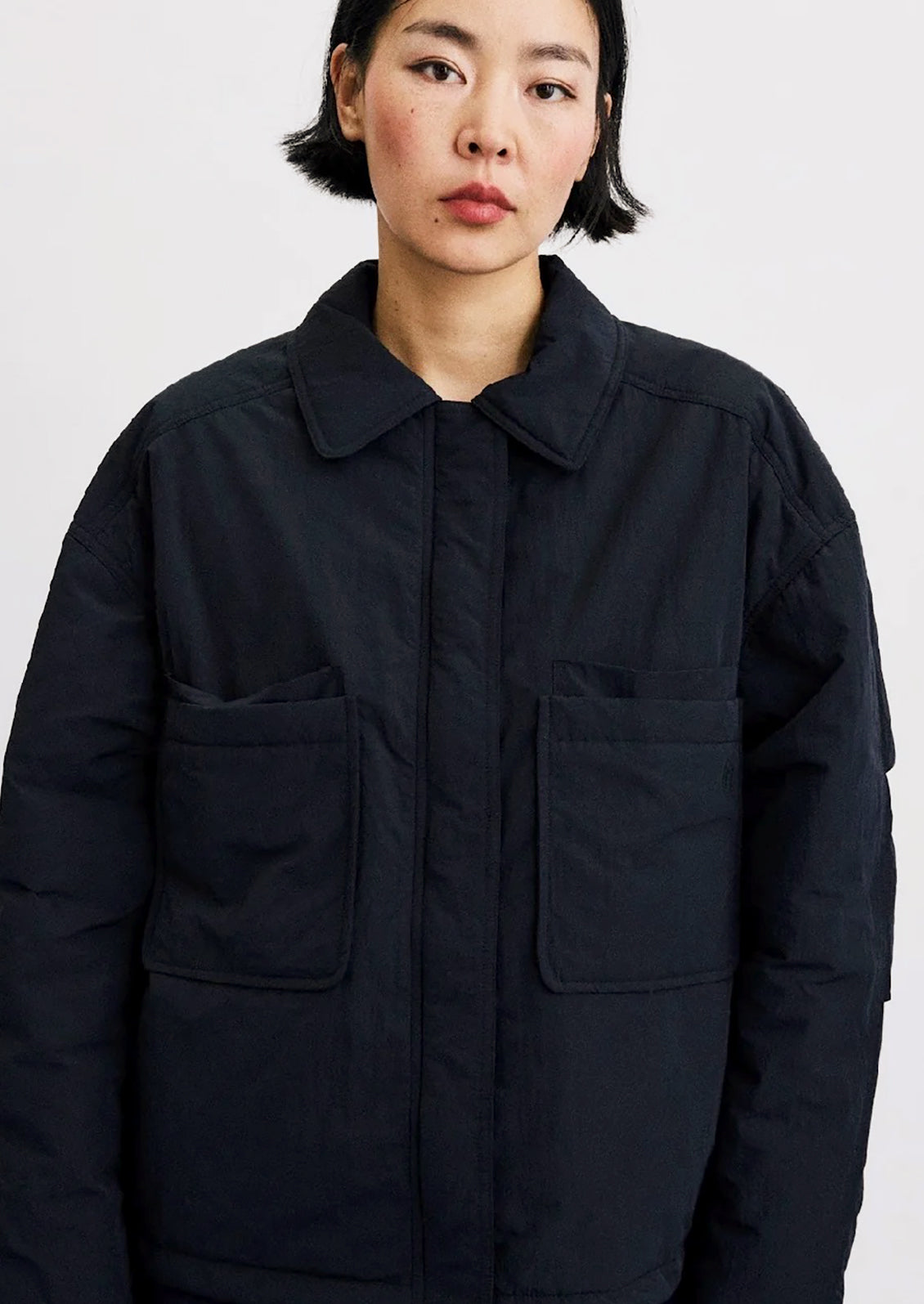A model wearing a dark navy blue utility jacket with a collar, front button closure, and two large patch pockets on a light gray background.