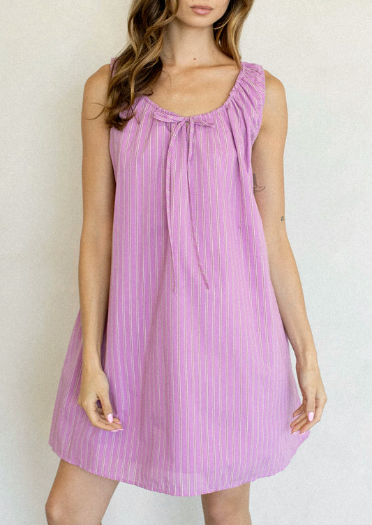 A woman wearing a sleeveless lavender-purple dress with thin vertical pink stripes and a tie-neck detail, standing against a plain light background.