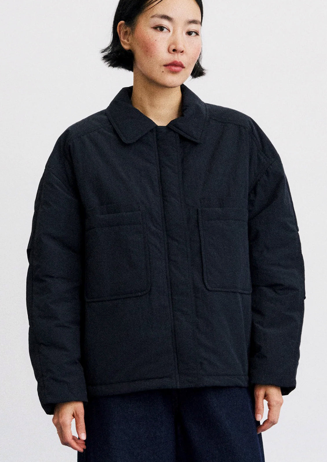 A model wearing a dark navy quilted jacket with a collar, front pockets, and a button closure, set against a plain light gray background.
