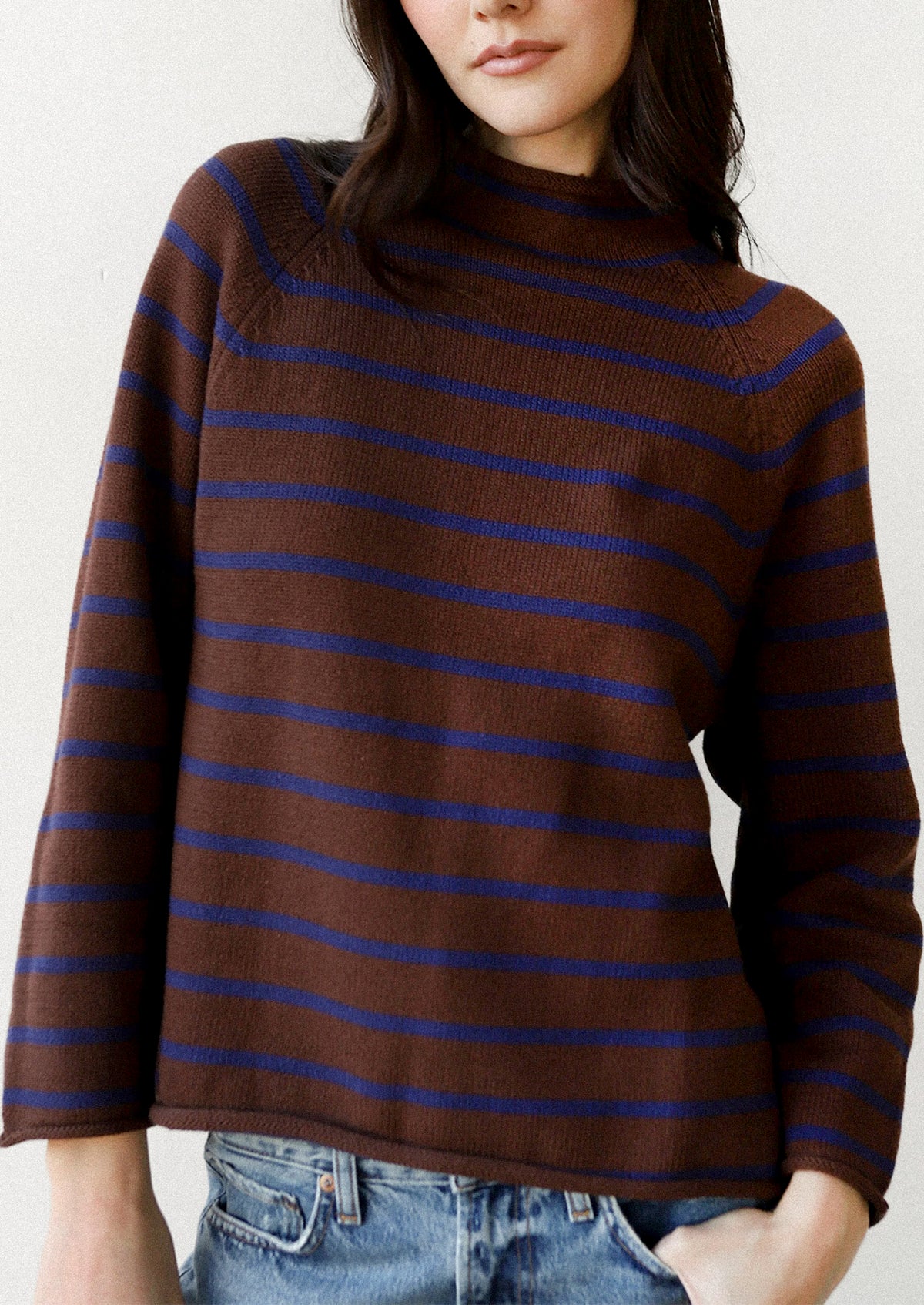 A woman wearing a brown knit mock-neck sweater with thin horizontal blue stripes, paired with blue jeans, shown against a plain off-white background.