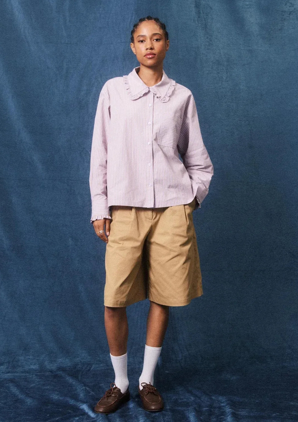 A model stands against a textured blue backdrop wearing a light pink long-sleeve button-up shirt with a ruffled collar, tan shorts, white socks, and brown loafers.
