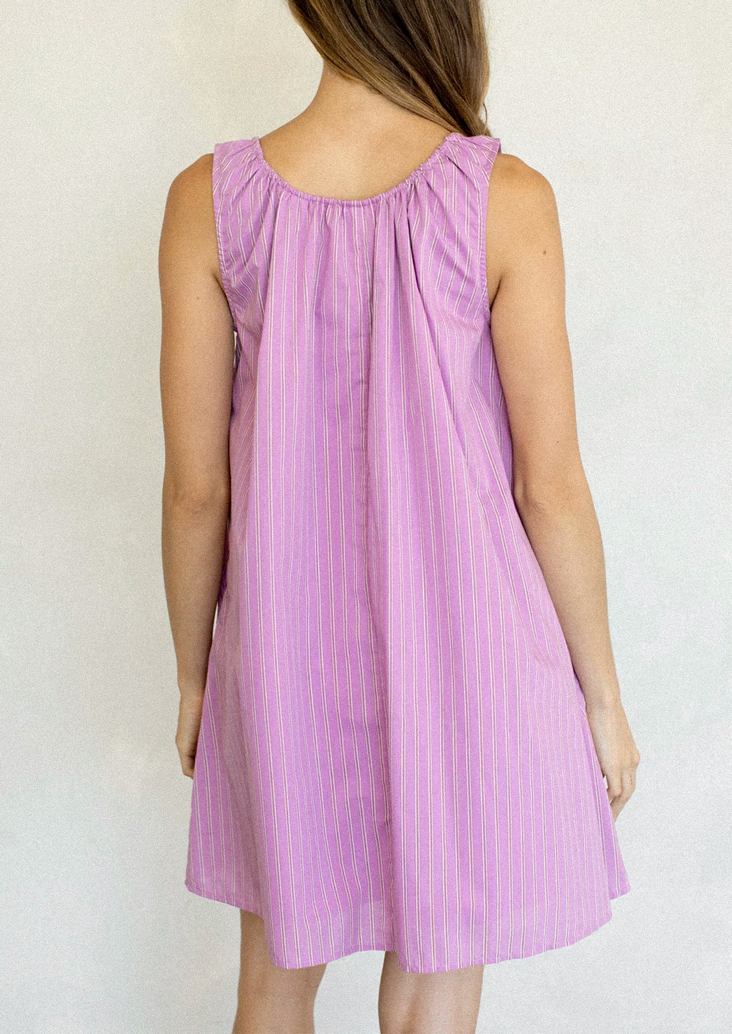 A woman wearing a sleeveless lavender-purple dress with thin vertical pink stripes and a tie-neck detail, standing against a plain light background.