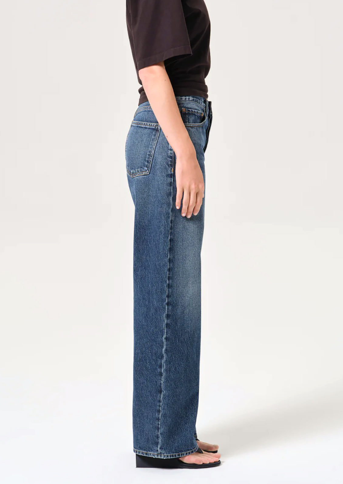 A side-profile view of a model wearing blue wide-leg denim jeans, a dark brown t-shirt, and black sandals against a plain white studio background.