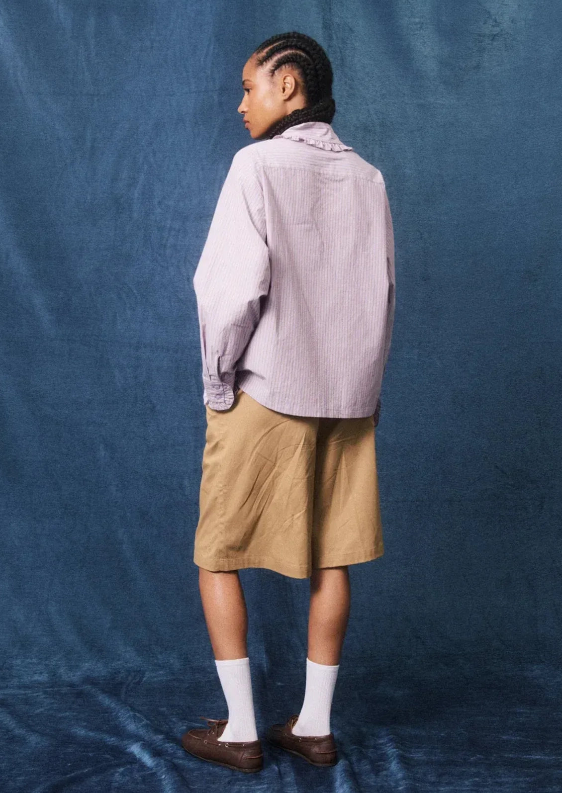 A model is shown from the back wearing a light lavender striped button-up shirt, tan shorts, white socks, and brown loafers, standing against a textured blue backdrop.