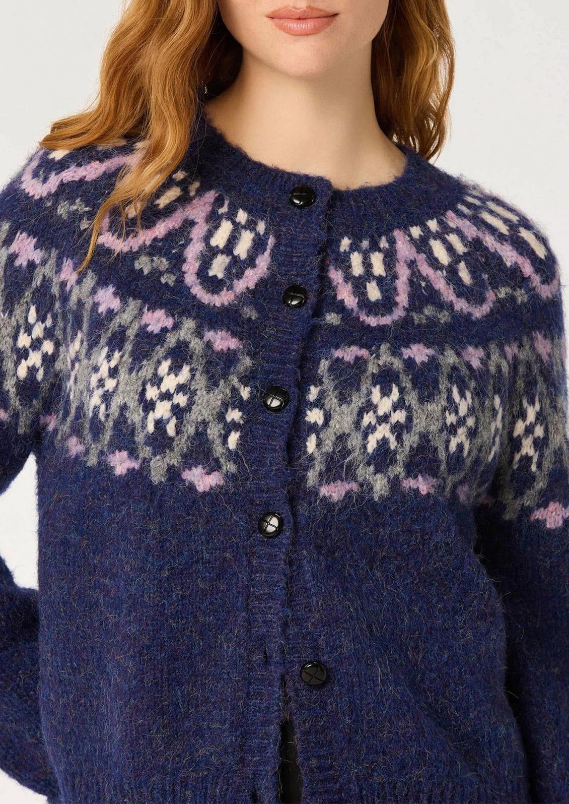 A close-up of a woman wearing a fuzzy knit cardigan sweater with a navy blue base and a Fair Isle style pattern in light pink, white, and gray. The cardigan features a round neckline and a front button closure.