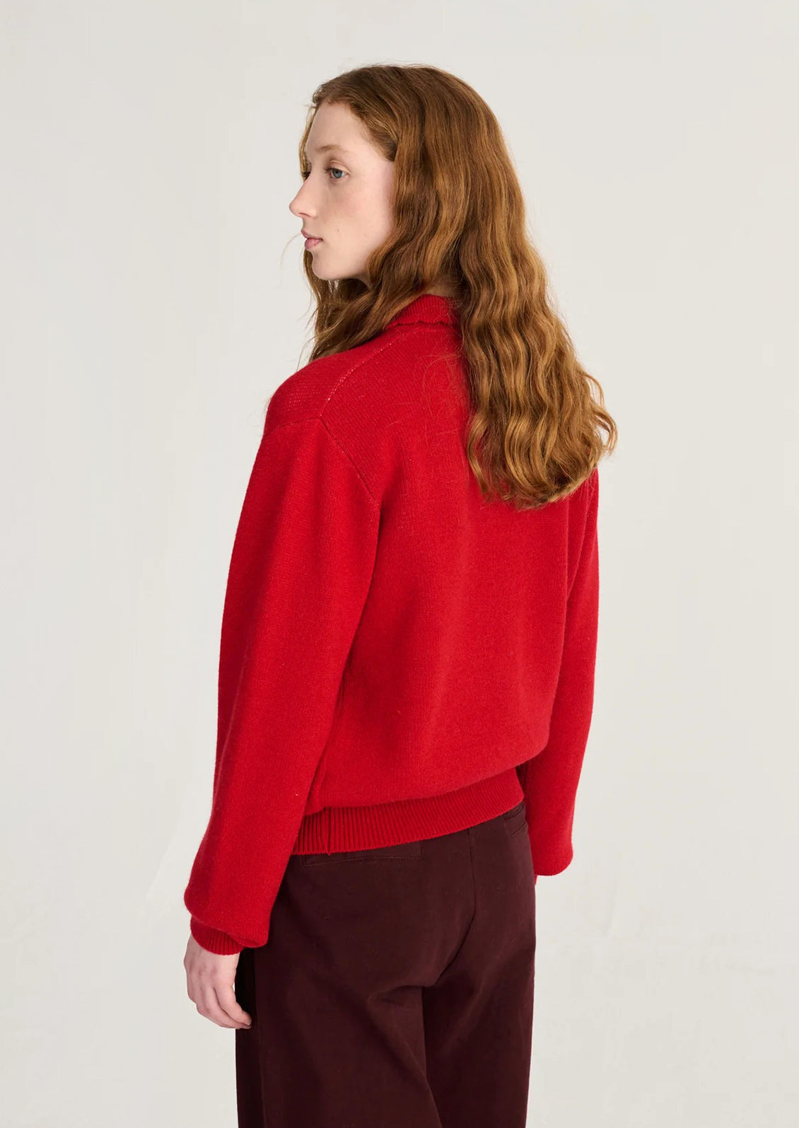 A woman with long wavy red hair is shown from a side-back angle wearing a bright red knit sweater and dark burgundy trousers against a plain light background.