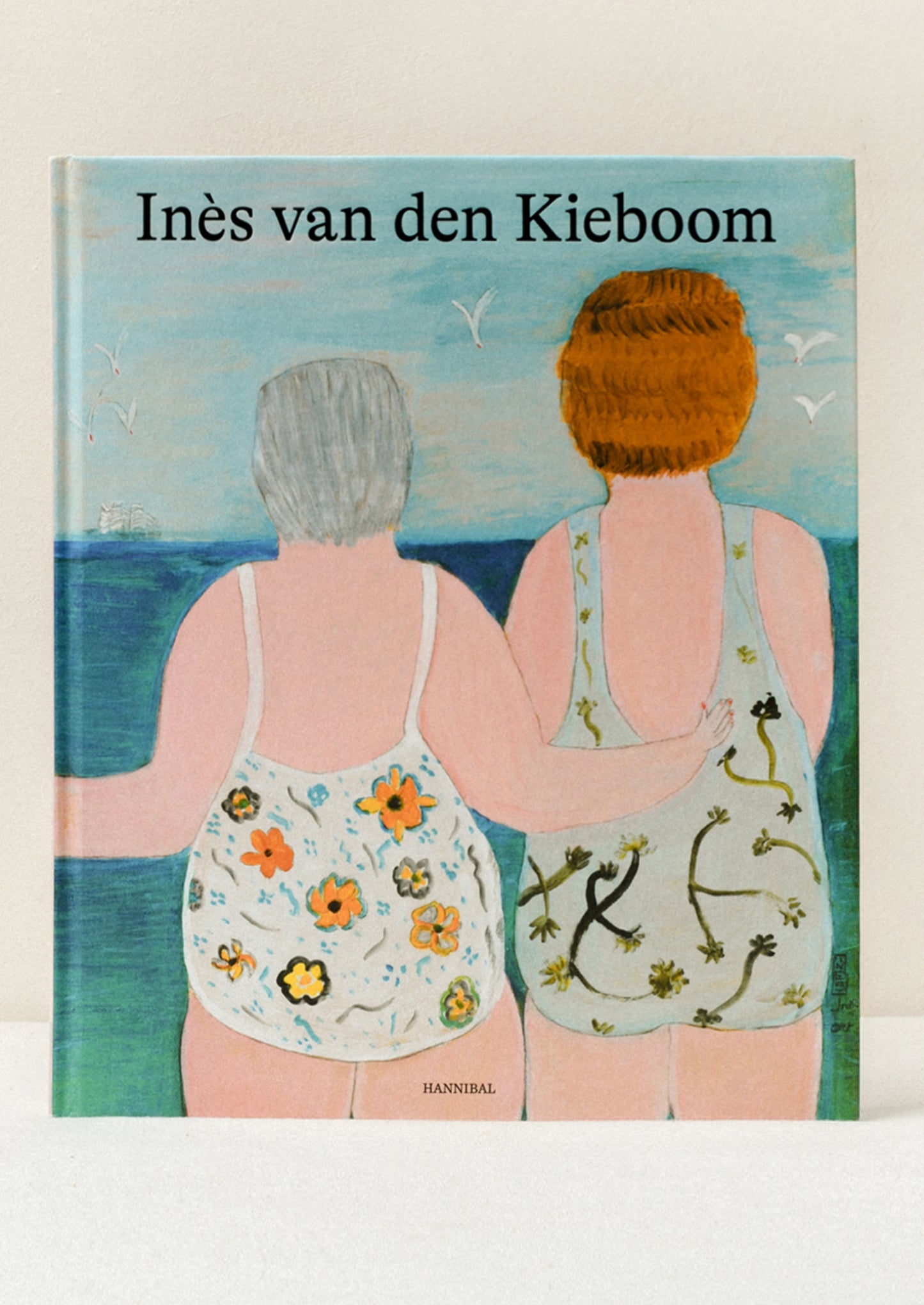 A hardcover book standing upright on a table titled "Ines van den Kieboom" featuring an illustration of two women in floral one-piece swimsuits facing the ocean with seagulls flying in the sky.