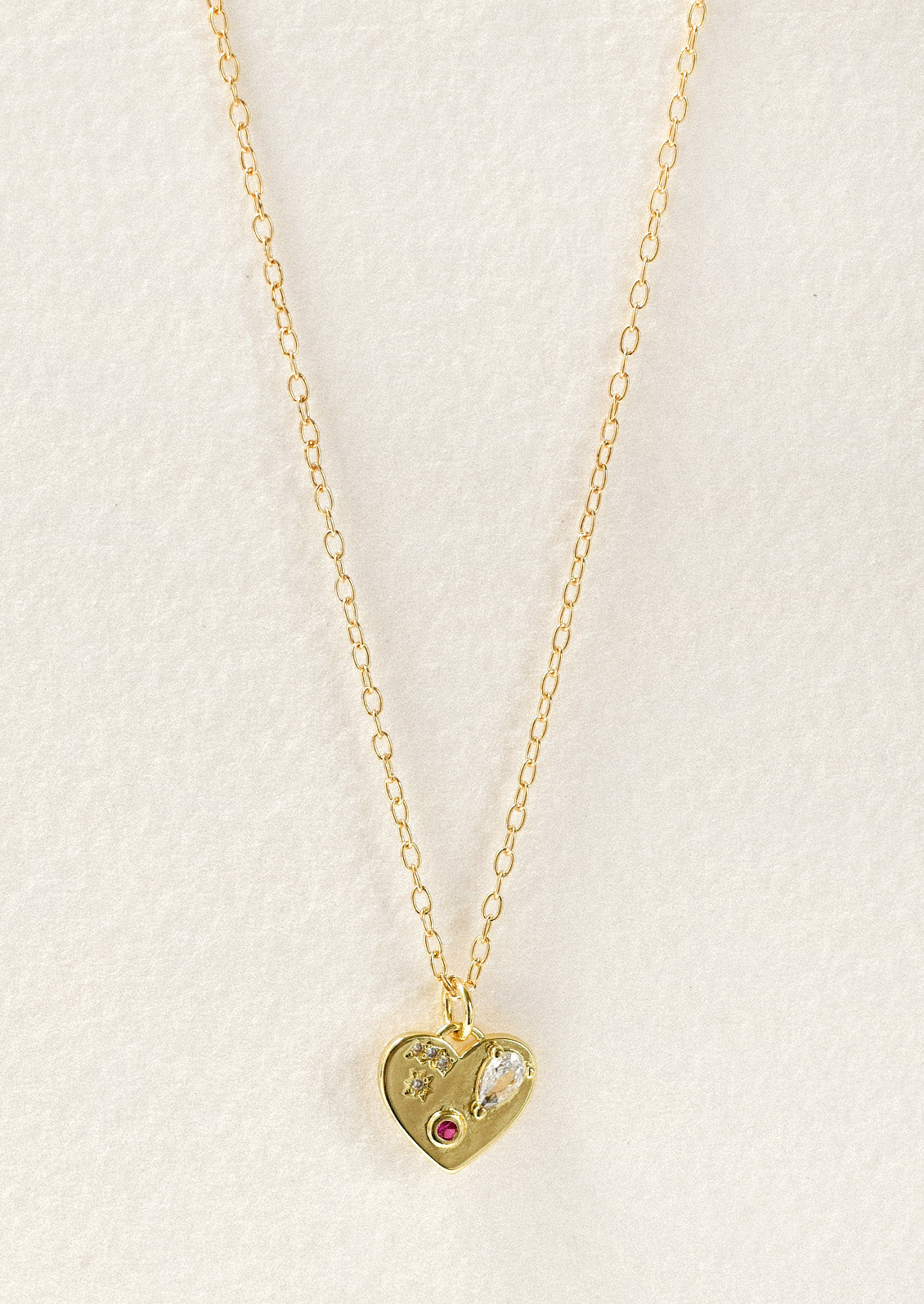 A gold necklace with a gold heart charm adorned with glass crystal and a red gemstone.