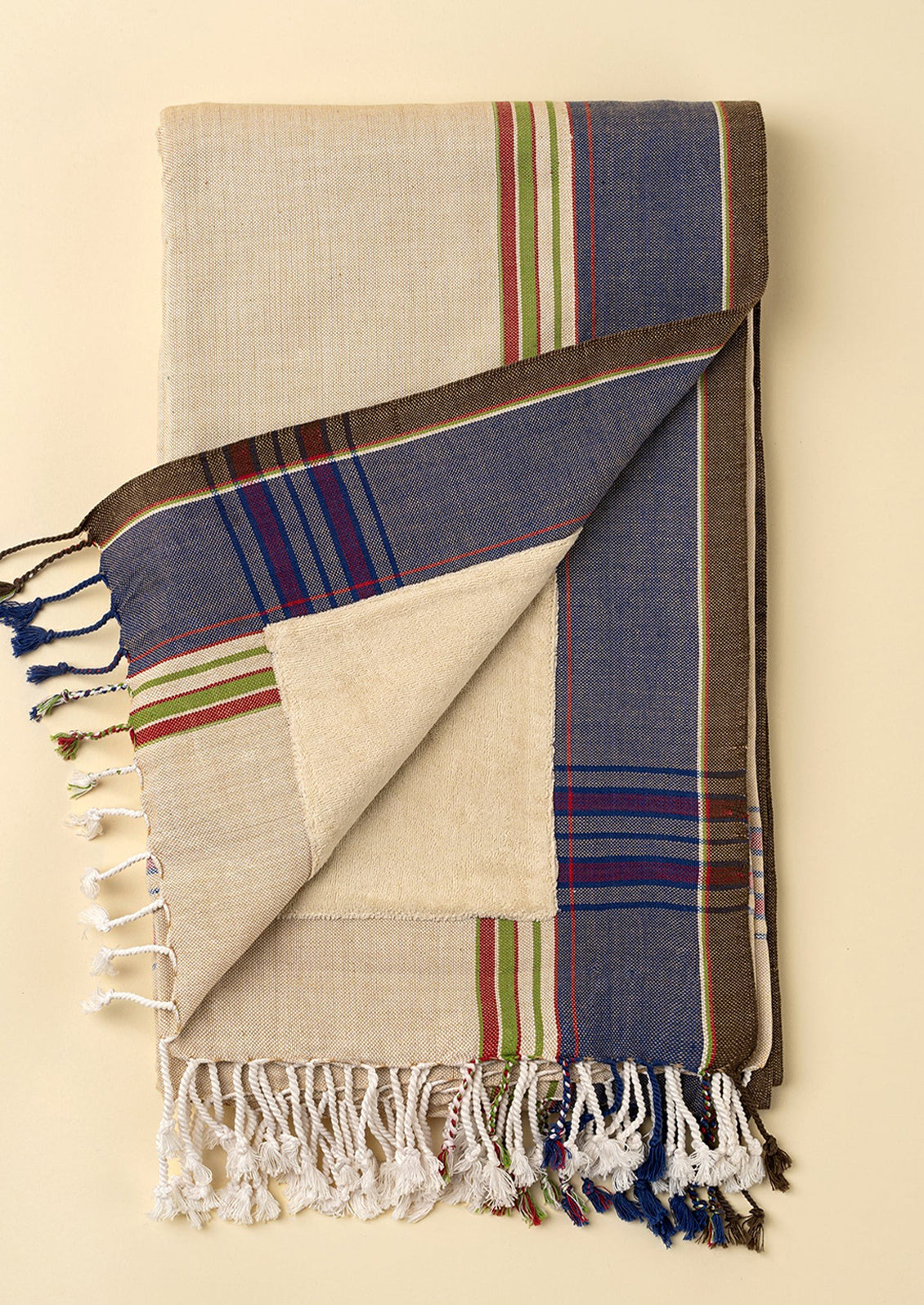 A folded woven throw blanket with fringed tassels, shown on a warm cream background. The blanket has a cream base with a blue border featuring red, green, and blue stripes, and a brown stripe running vertically.