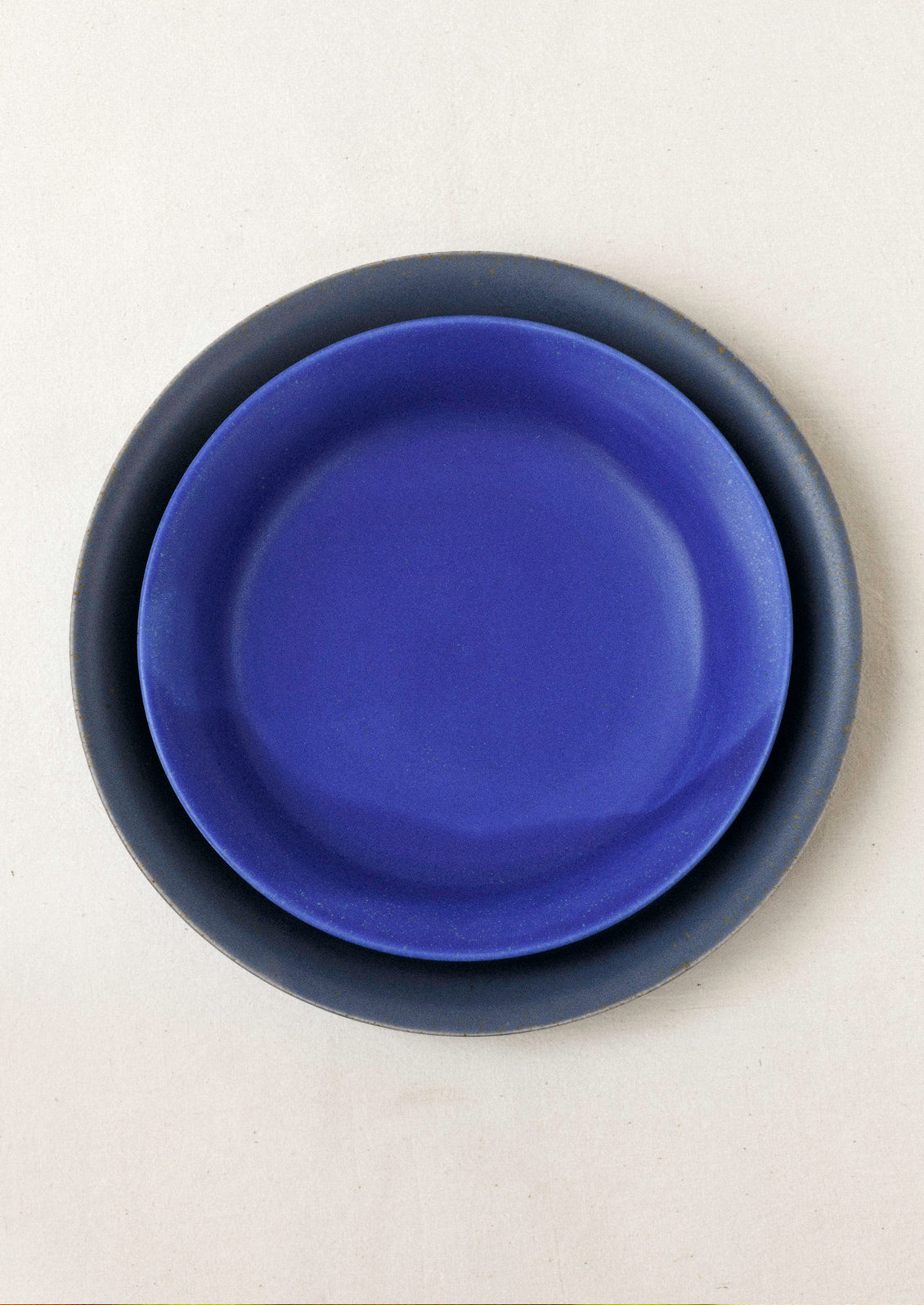 A large dark grey ceramic dinner plate with a smaller cobalt blue ceramic dinner plate on top set on a cream background.