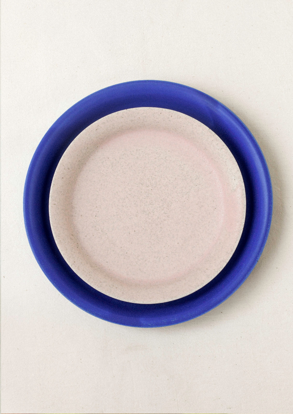 A large cobalt blue ceramic dinner plate with a smaller light pink ceramic dinner plate on top set on a cream background.