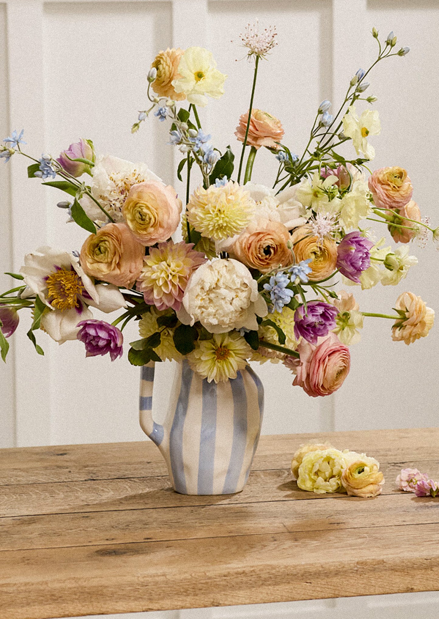 A large, vibrant floral arrangement featuring a mix of pastel yellow, peach, cream, purple, and blue flowers, presented in a blue and white vertically striped ceramic pitcher on a rustic wooden table.