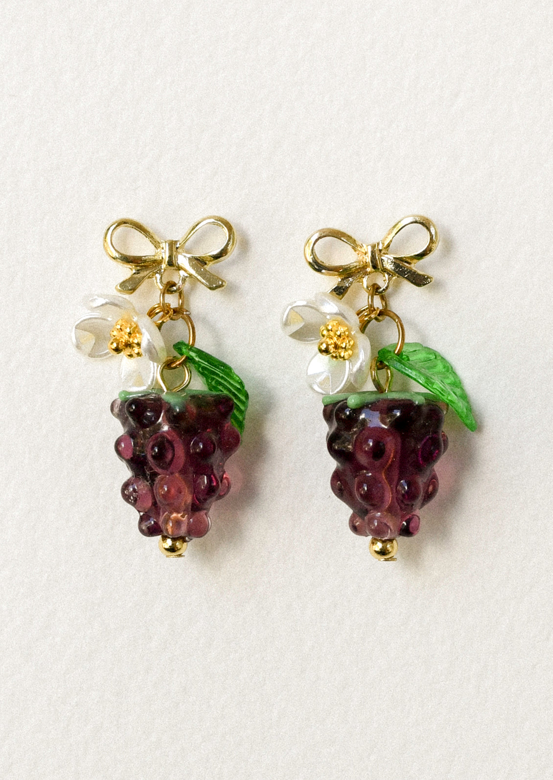 A pair of gold bow stud earrings with dangling glass blackberries along with a white flower and green leaf.
