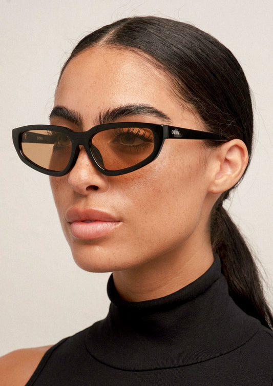 A woman wearing a pair of angular cat eye sunglasses with a black frame and light brown lenses.