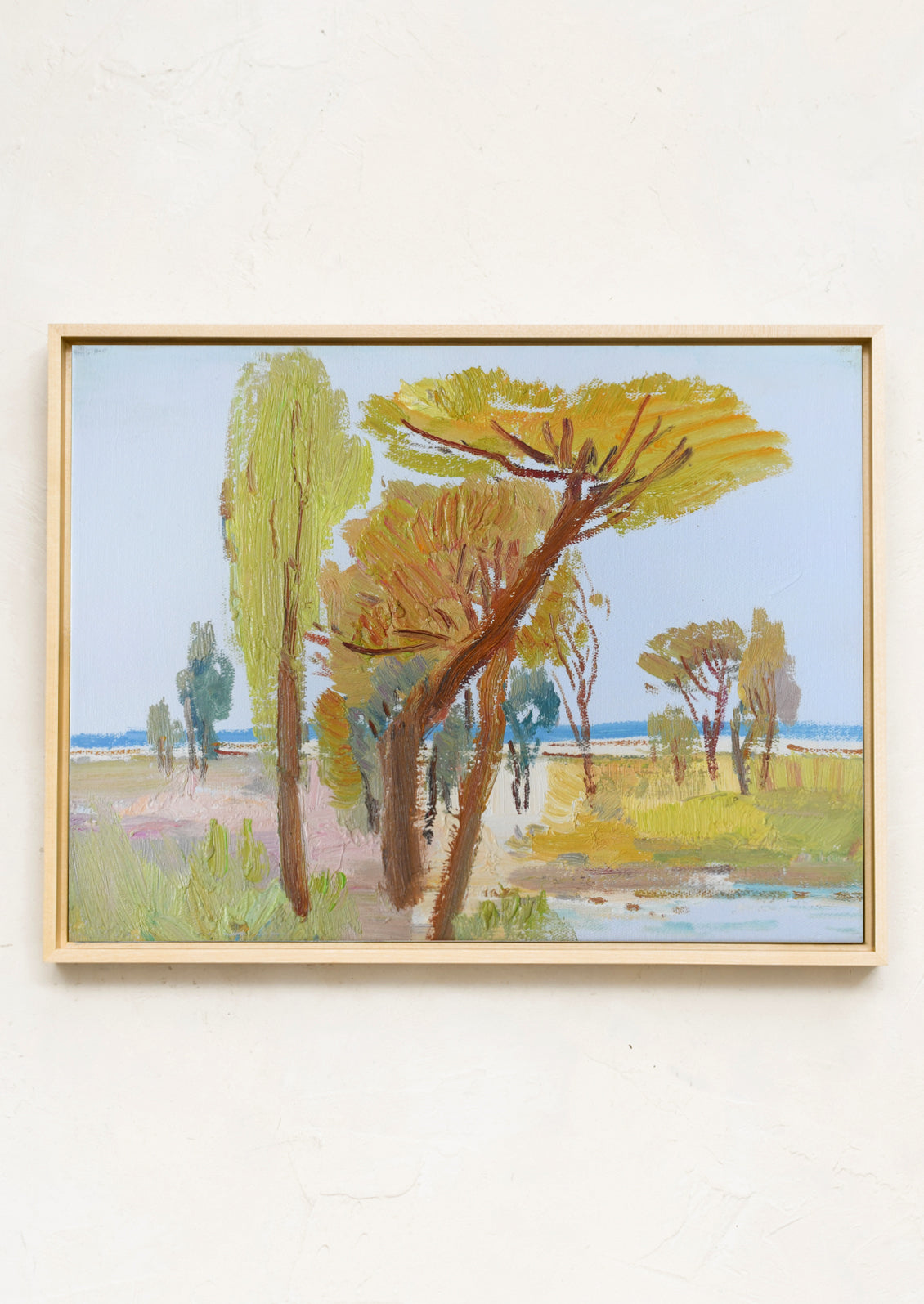 A framed landscape painting featuring tall trees in shades of green, yellow, and brown against a pale blue sky, hung on a textured off-white wall.