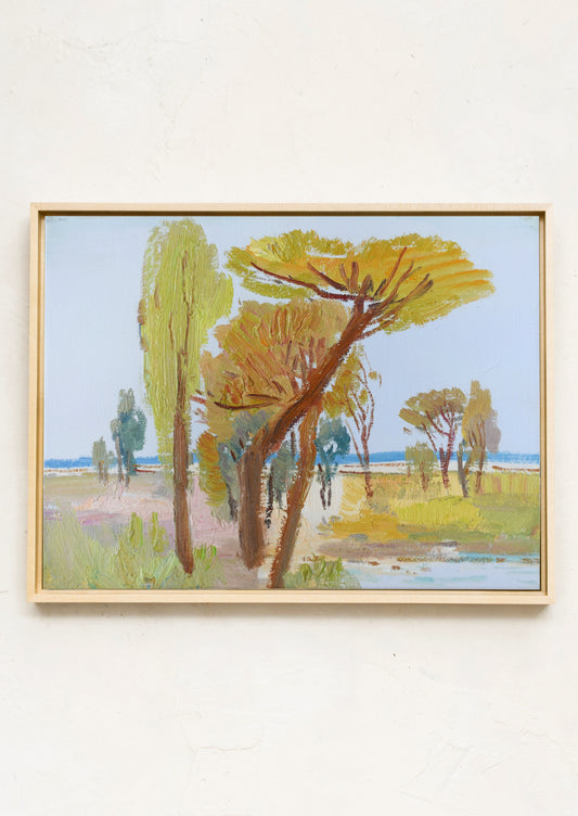 A framed landscape painting featuring tall trees in shades of green, yellow, and brown against a pale blue sky, hung on a textured off-white wall.