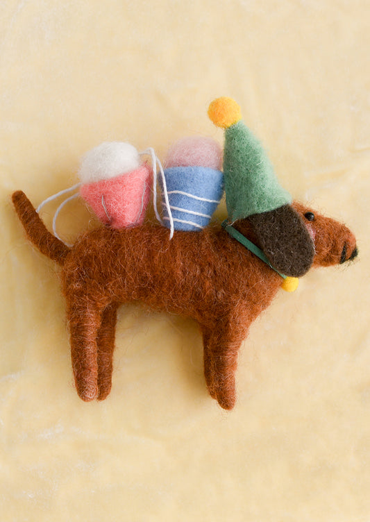 A felted ornament of a brown dog wearing a party hat and carrying cupcakes on its back.