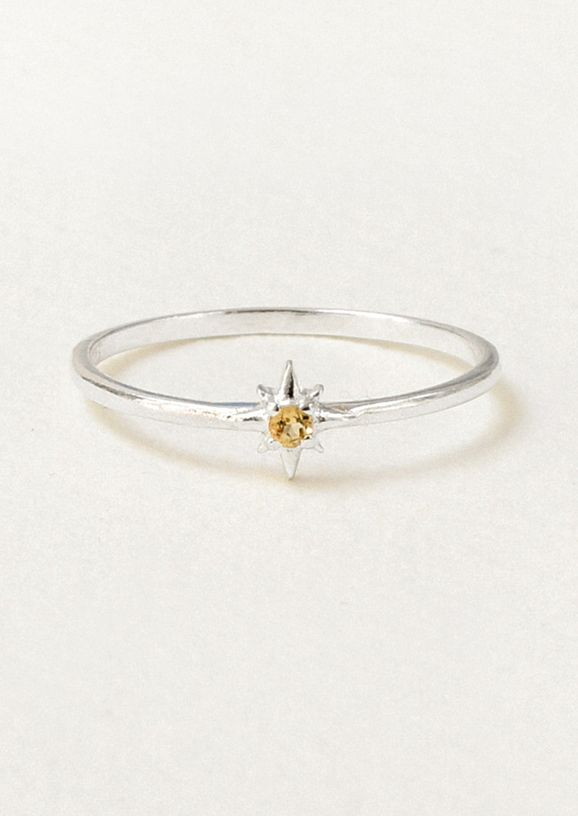 A delicate sterling silver ring featuring a starburst setting with a small round yellow gemstone, photographed on a clean, light beige background.