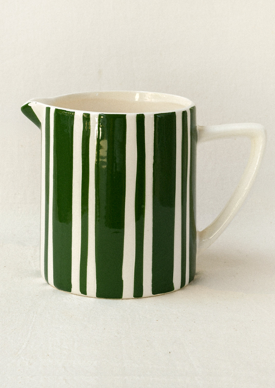 A ceramic pitcher with a handle and spout, decorated with vertical dark green and white stripes, set against a plain off-white background.