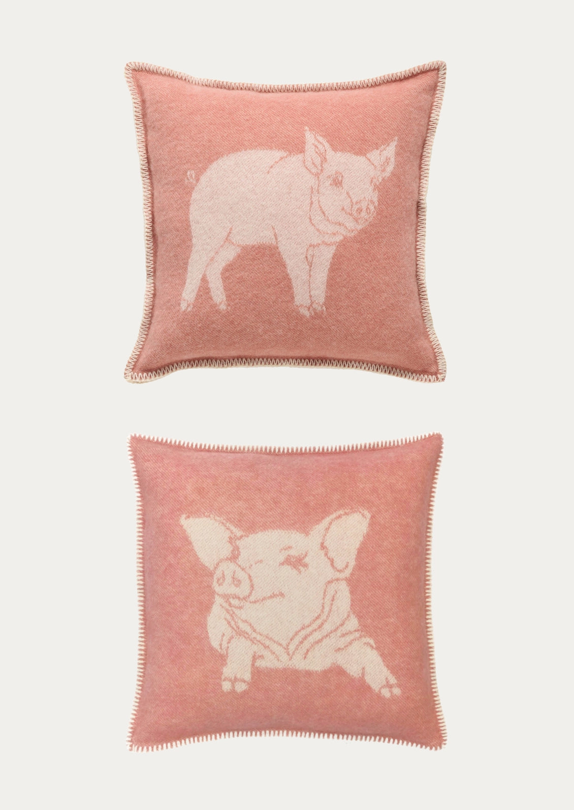 Two square pink throw pillows with tufted pom-pom trim, featuring knitted pig illustrations in contrasting light pink tones, shown against a light gray background.