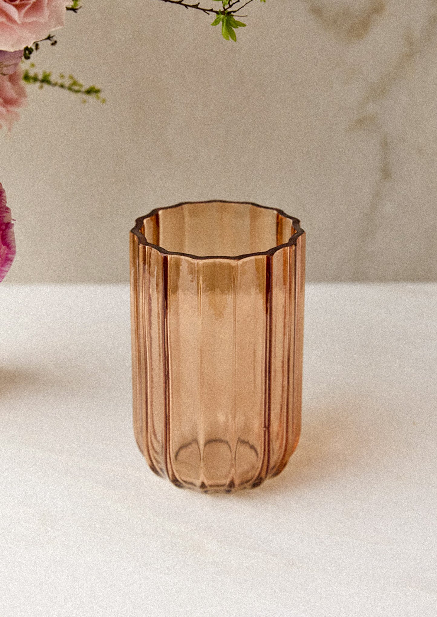 A ribbed, translucent glass vase with a scalloped rim, set on a light marble surface with soft pink flowers in the background.