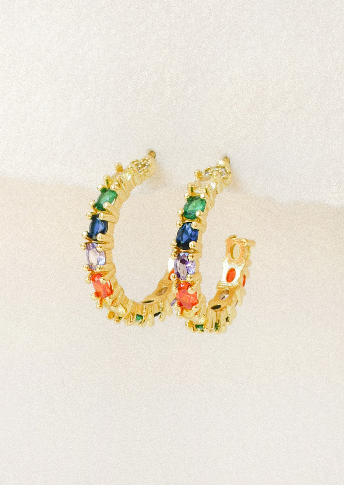 A pair of gold hoop earrings adorned with various colored gems hanging from a white paper.