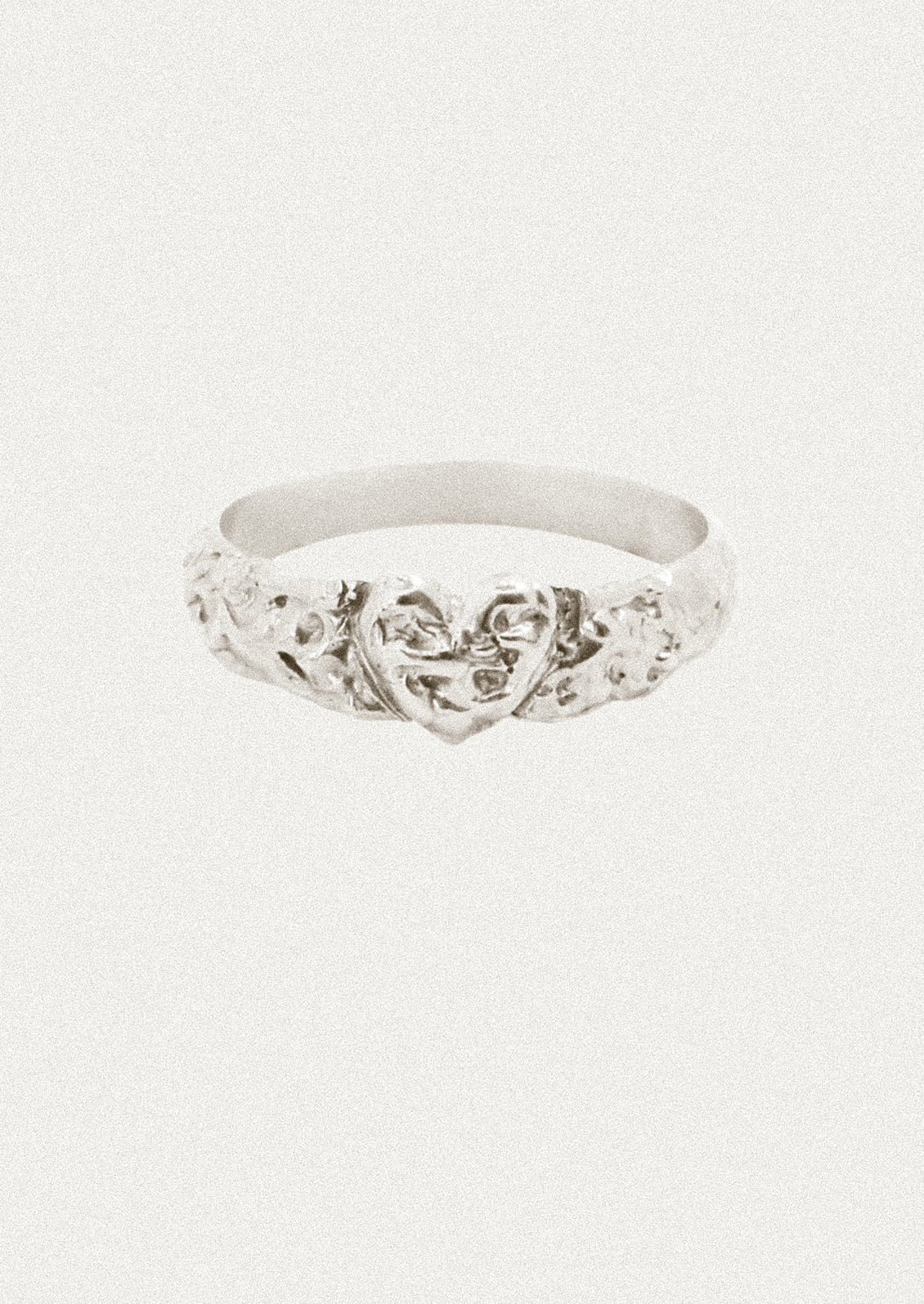 A silver band ring featuring a central heart motif surrounded by delicate floral and vine details, set against a plain off-white background.