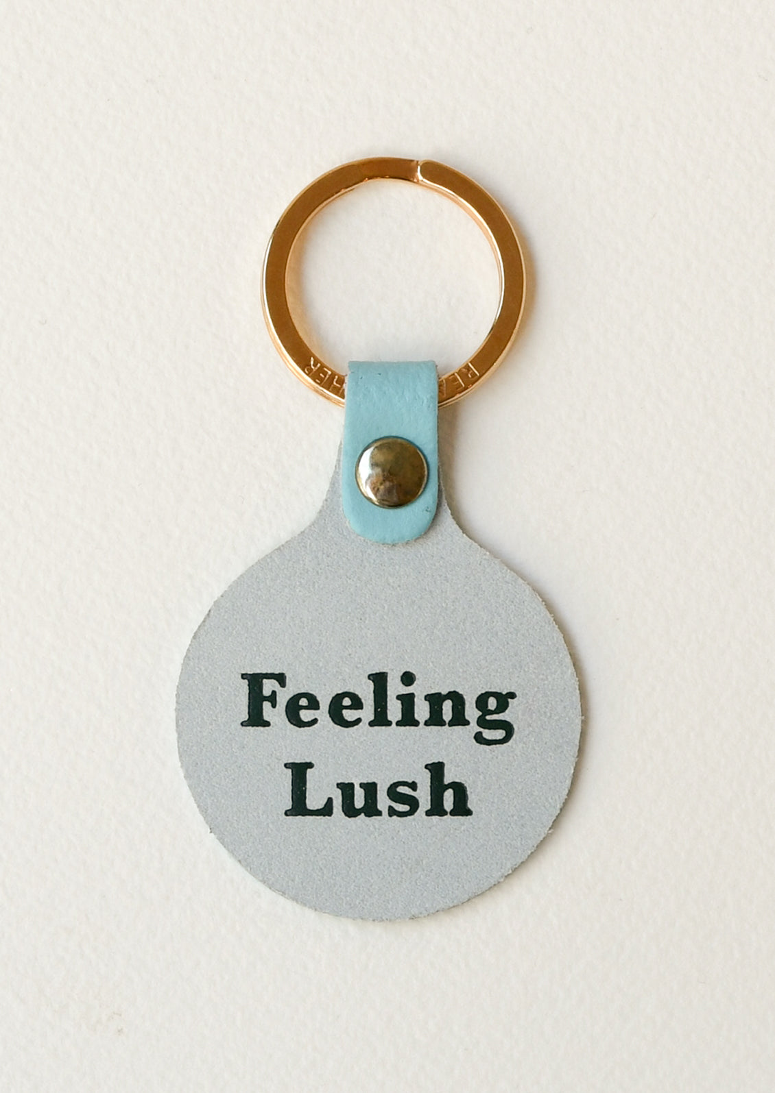 The back of a turquoise leather circle keychain with the words "Feeling Lush" on the back on a gold key ring.