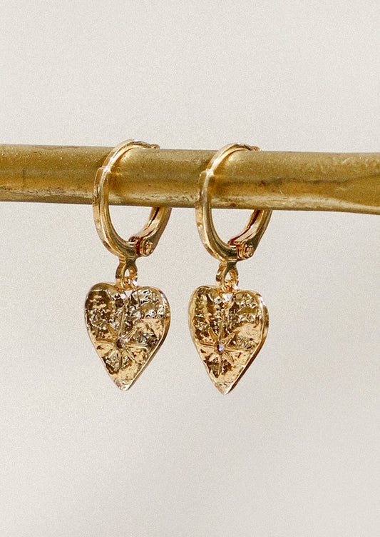 A pair of gold huggie hoop earrings with heart charms with a starburst design hanging from a gold pole in front of a gray background.