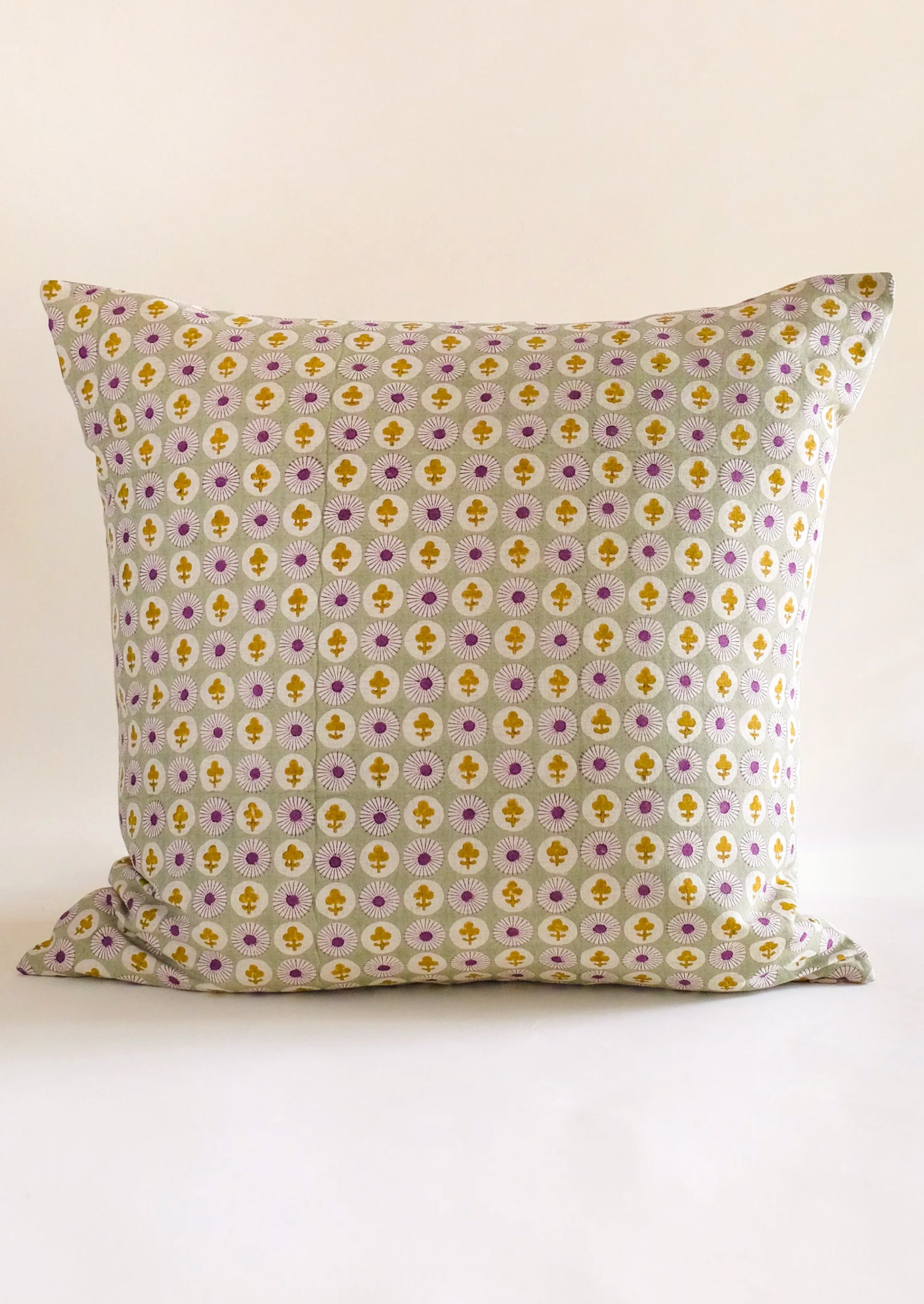 A square throw pillow with a repeating geometric pattern in yellow, purple, and white on a light gray background, photographed against a plain off-white backdrop.