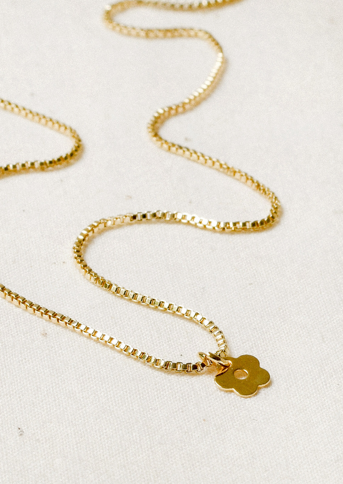 A gold chain necklace with a tiny daisy charm laying on a white background.