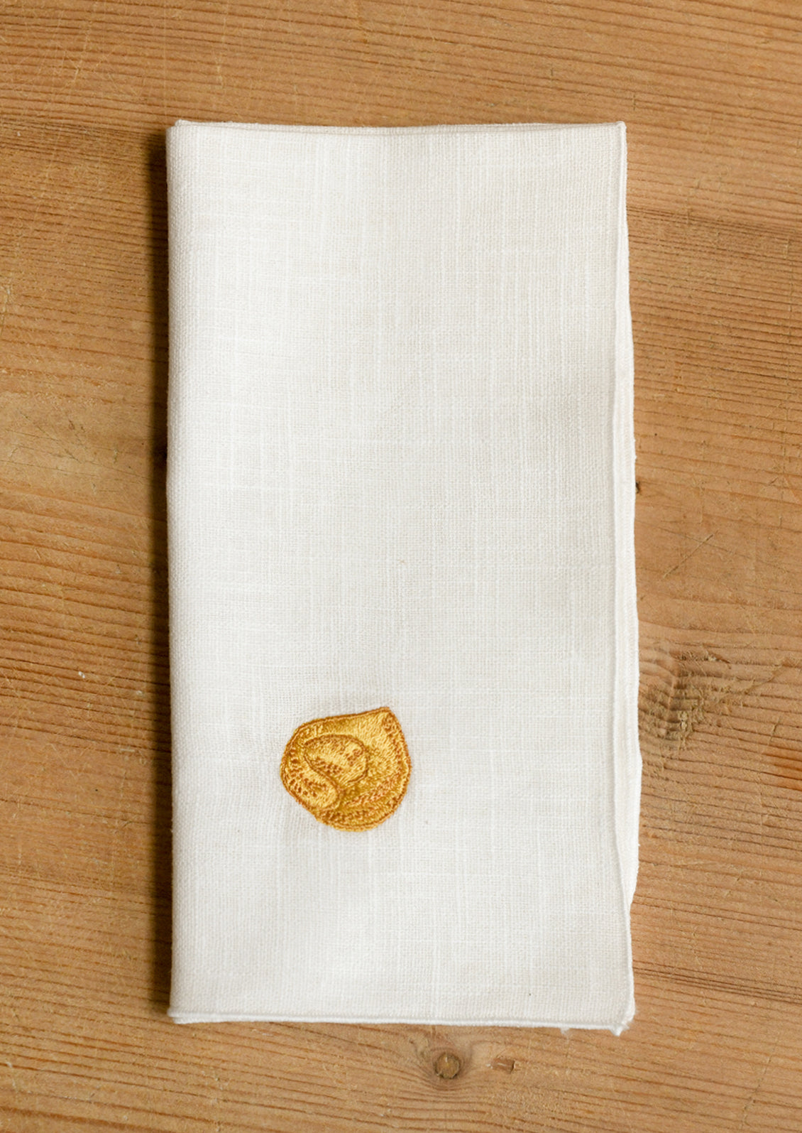 A folded off-white linen napkin with a small yellow embroidered tortellini, displayed on a light brown wooden surface.