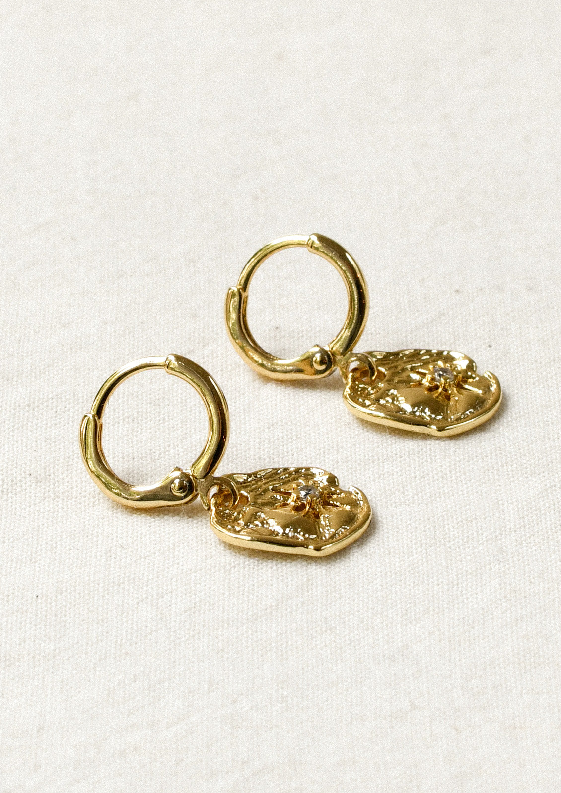 A pair of gold huggie hoop earrings with dangling disc charms that have a starburst design and a cubic zirconia in the center.
