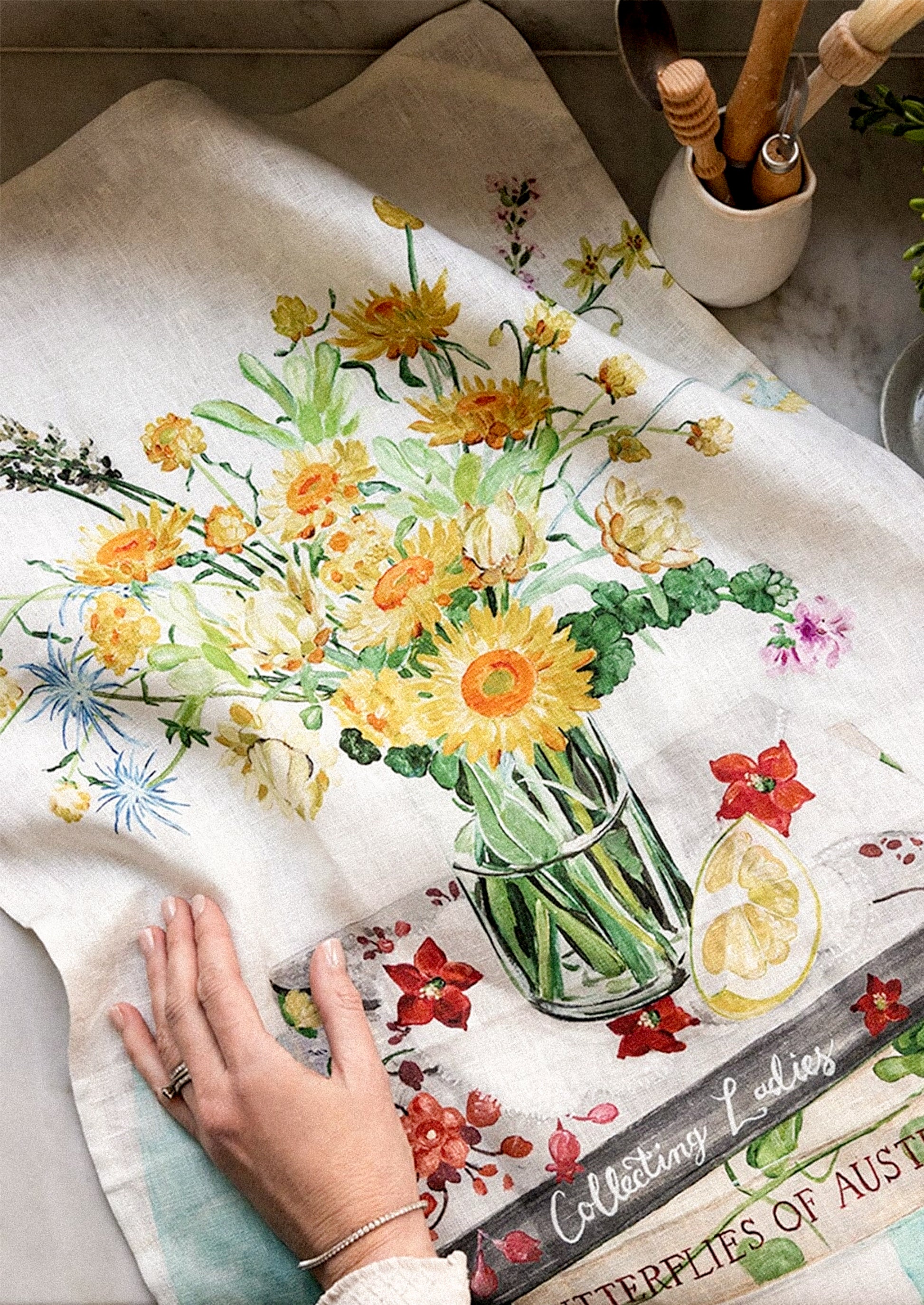 A linen tea towel with a colorful watercolor floral print of sunflowers and wildflowers in a glass vase.