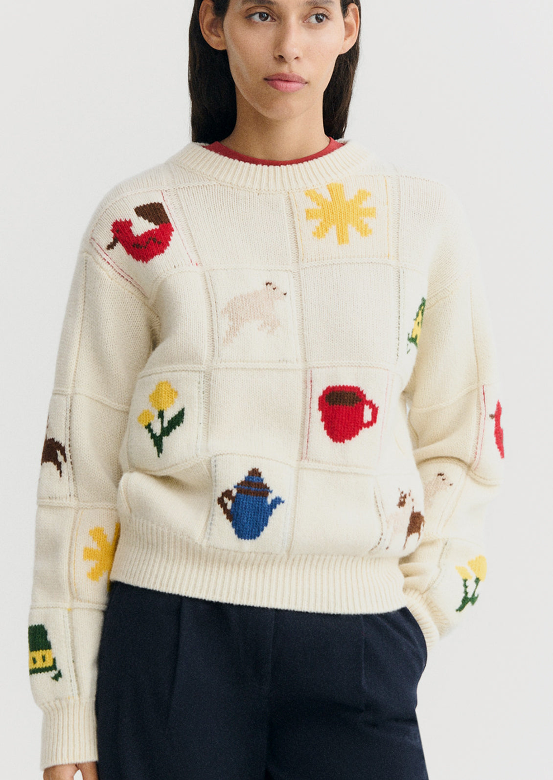 A woman wearing a cream-colored knit sweater with a patchwork design featuring colorful motifs like flowers, teapots, a red mug, and a dog, paired with dark navy trousers against a light gray background.