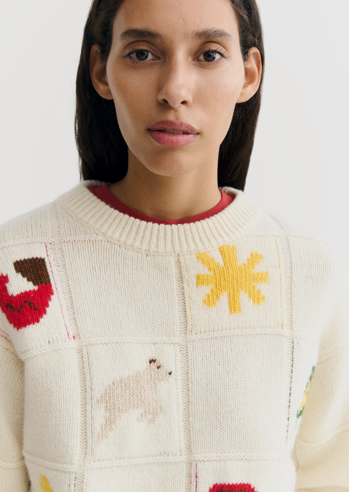 A close-up of a woman wearing a cream-colored knit sweater with colorful patchwork motifs, including a yellow sun, a red-and-brown bird, and a pale polar bear.