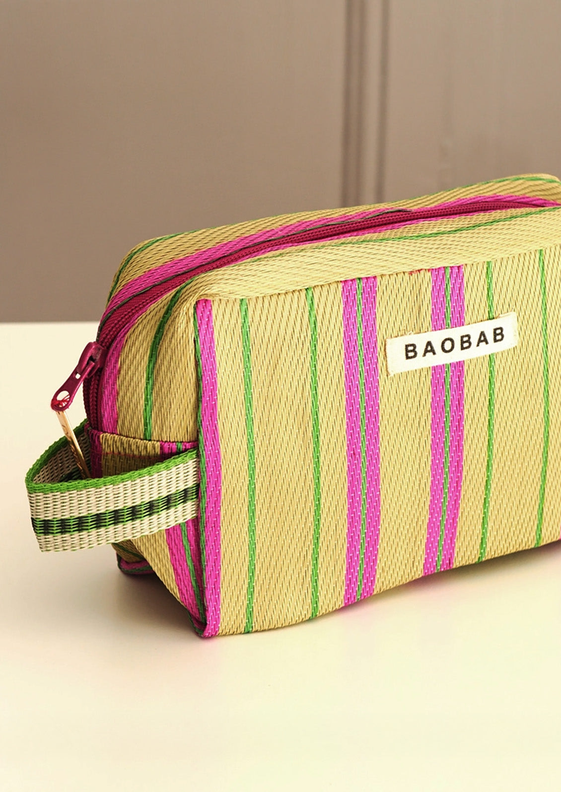A woven rectangular zip pouch in beige with vertical pink and green stripes, featuring a small white label that reads BAOBAB and a matching striped handle, photographed on a light surface with a neutral background.