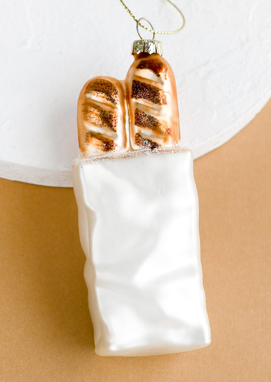 An all glass holiday ornament of a white paper bag with two baguettes poking out of the top.