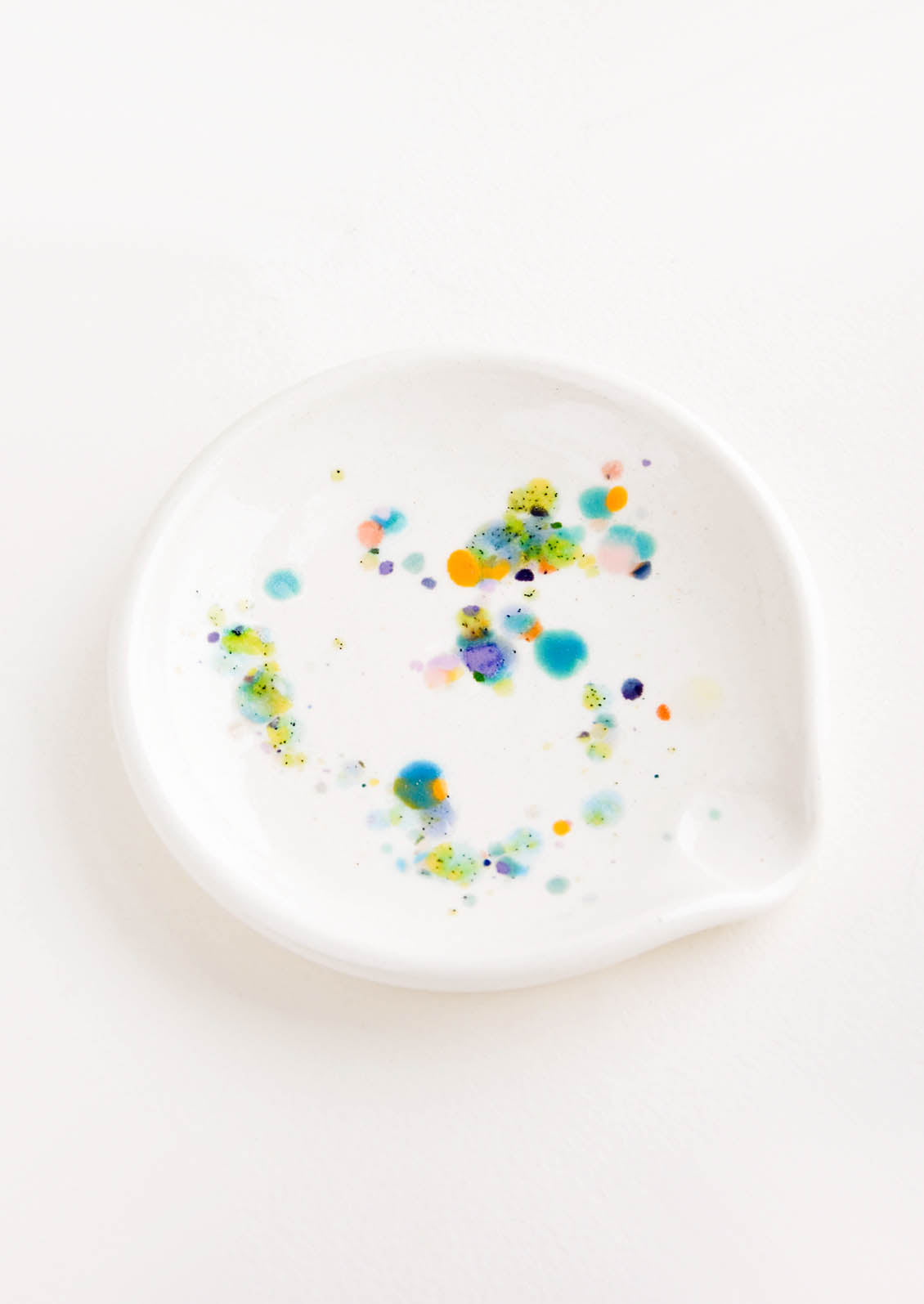 A round ceramic spoon rest with random, colorful glaze drips.