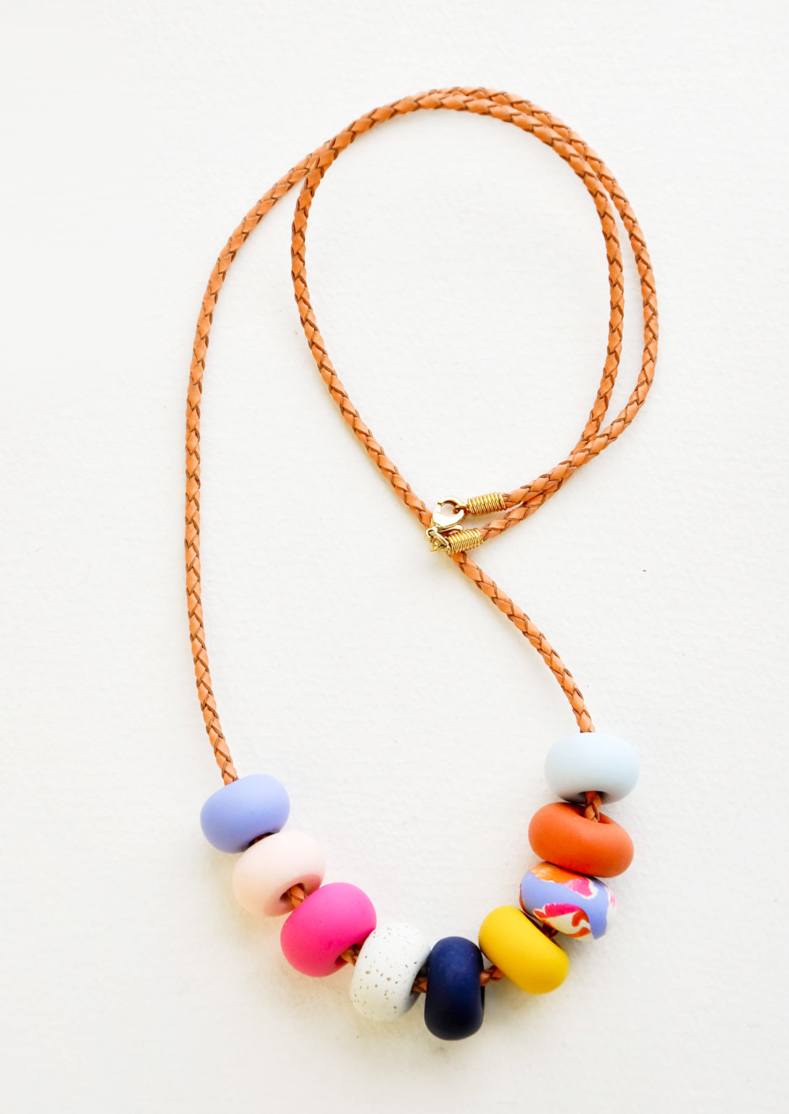 Necklace with gold clasp, brown woven leather cord, and nine multi-colored clay beads in blues, pinks, yellows, and oranges. 