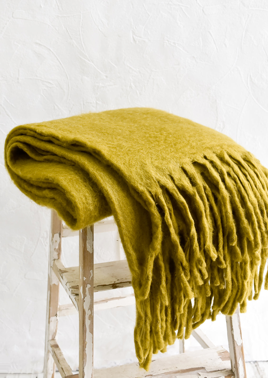 A chartreuse mohair blanket with long tassel fringe, folded on a stool.